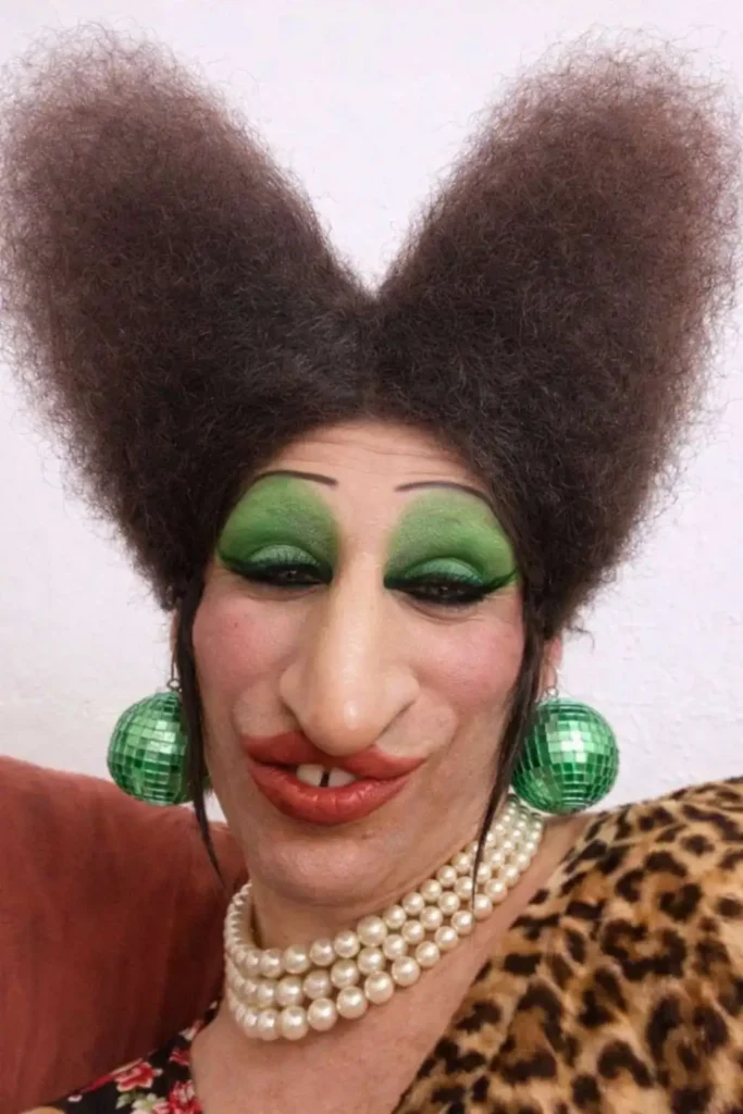 Person wearing a voluminous bow-shaped afro hairstyle paired with bold green eye makeup, large earrings, and a dramatic costume look