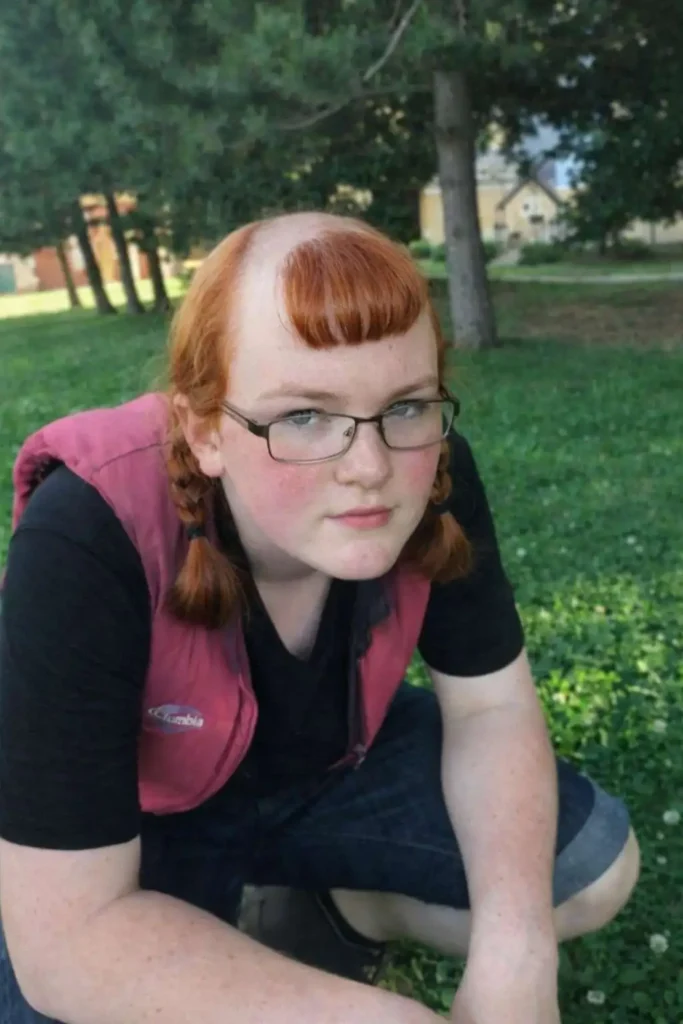 Person with a half-shaved haircut featuring a straight fringe and short pigtails, creating a bold and unconventional asymmetrical hairstyle